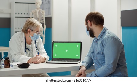 Doctor And Patient Looking At Laptop With Greenscreen At Chekcup Visit In Cabinet. Physician And Man Using Blank Copyspace Mockup With Isolated Chromakey Template During Covid 19 Pandemic.