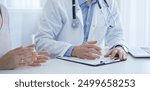 A doctor and a patient. The physician, wearing a white medical coat over a blue shirt, is filling out a medical record form during a consultation in the clinic. Medical service