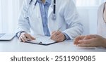 A doctor and a patient. The physician, wearing a white medical coat over a blue shirt, is filling out a medical record form during a consultation in the clinic. Medical service