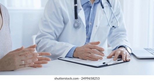 A doctor and a patient. The physician, wearing a white medical coat over a blue shirt, is filling out a medical record form during a consultation in the clinic. Medical service - Powered by Shutterstock