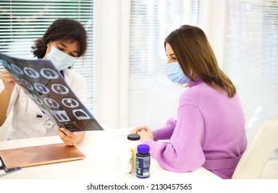 Doctor And Patient In Protective Face Masks Discuss X-ray Result During Appointment. African American Woman Physician With CT Lung Screening Explaining Diagnosis To Sick Woman. Treatments For COVID-19