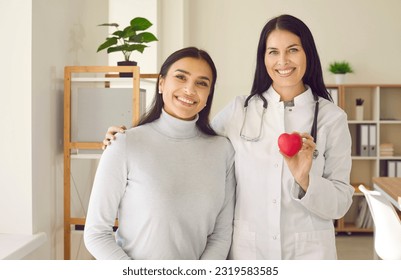 Doctor and patient reminding of importance of heart health and regular routine check ups. Happy healthy woman doctor and patient standing in clinic, holding red heart, looking at camera and smiling - Powered by Shutterstock
