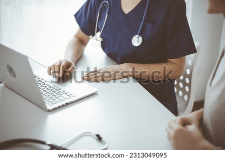 Doctor and patient sitting at the desk in clinic office. The focus is on unknown female physician's hands using a laptop computer, close up. Medicine concept