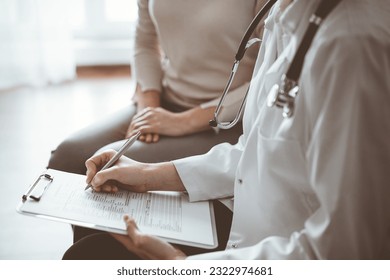 Doctor and patient sitting at sofa in clinic office. The focus is on female physician's hands filling up the medication history record form, close up. Medicine concept - Shutterstock ID 2322974681