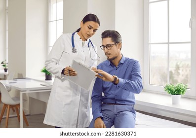 Doctor and patient talking during medical checkup at clinic. Young woman in white coat showing and explaining analysis results to man sitting on medical couch in examination room - Shutterstock ID 2281630371