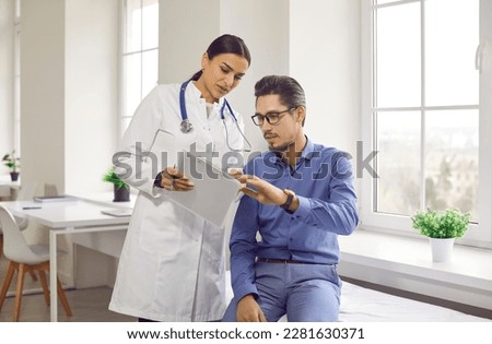 Doctor and patient talking during medical checkup at clinic. Young woman in white coat showing and explaining analysis results to man sitting on medical couch in examination room