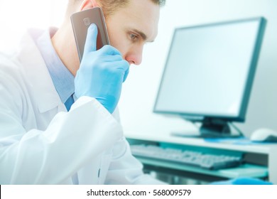 Doctor Phone Call Consultation. Medical Doctor Using His Phone And Computer To Get Second Opinion. Mid 30s Caucasian Doctor.