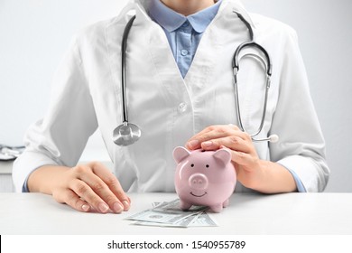 Doctor with piggybank and money at white table indoors, closeup. Medical insurance concept