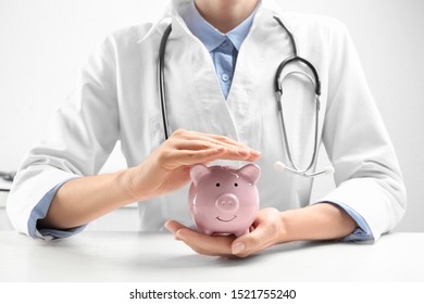 Doctor with piggybank at white table indoors, closeup. Medical insurance concept