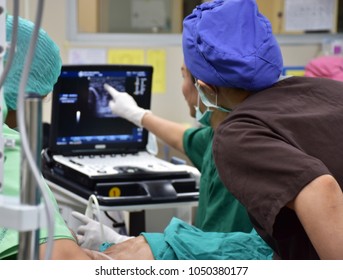 Doctor Is Pointing Finger To Teach Intern To Use Ultrasound For Fine Vessel For Approach Pressure Line In Operating Room. Focus At Intern, Blurred Doctor And Ultrasound Machine.