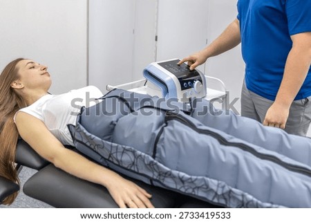 A doctor points at the device screen while a woman receives pressotherapy on a treatment table in a rehabilitation center. Tech focused look, clean interior, recovery and selfcare trend.