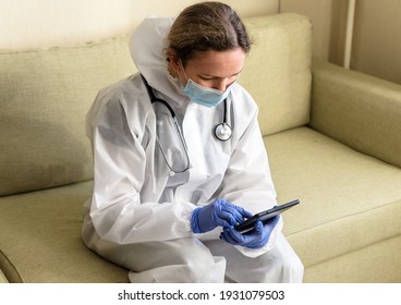 Doctor In PPE Suit Uses Digital Tablet In COVID-19 Patient Home, Female Physician In Coverall And Mask Looks At Gadget During COVID Coronavirus Pandemic. Concept Of Corona Virus Treatment And Care.