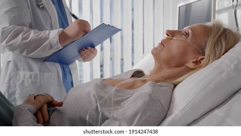 Doctor In Professional Uniform Examining Patient At Hospital Or Medical Clinic. Sick Woman With Nasal Oxygen Catheter Lying In Bed And Talking To Doctor In Hospital Ward