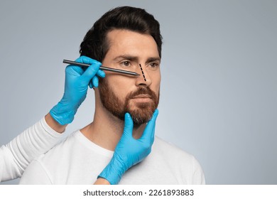 Doctor in protective gloves drawing marks on man's nose for cosmetic surgery operation, standing against grey studio background, free space