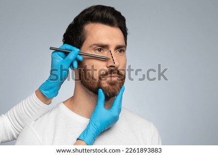 Doctor in protective gloves drawing marks on man's nose for cosmetic surgery operation, standing against grey studio background, free space
