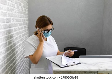 A Doctor In A Protective Mask Speaks On The Phone With A Patient And Reviews The Records At The Reception Desk In The Clinic. Make An Appointment By Phone