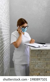 A Doctor In A Protective Mask Speaks On The Phone With A Patient And Reviews The Records At The Reception Desk In The Clinic. Make An Appointment By Phone