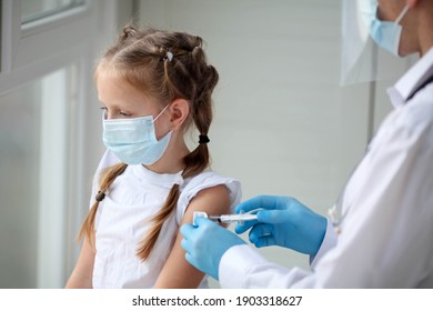 A Doctor In A Protective Suit Makes The Child  In A Medical Mask A Shoulder Shot . Coronavirus Vaccination. Covid-19 Vaccine. Doctor Vaccinating Child. Kids At Clinic. Little Girl Getting Flu Shot. 
