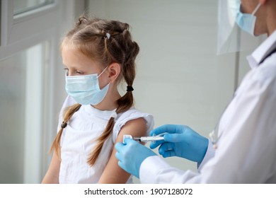 A Doctor In A Protective Suit Makes The Child  In A Medical Mask A Shoulder Shot . Coronavirus Vaccination. Covid-19 Vaccine. Doctor Vaccinating Child. Kids At Clinic. Little Girl Getting Flu Shot. 