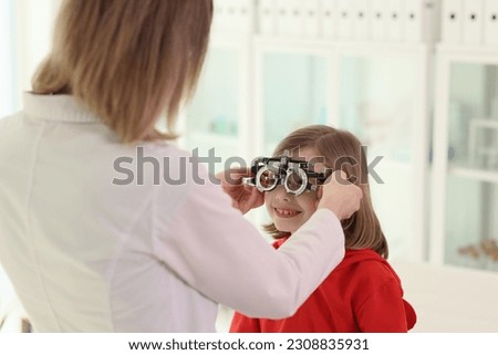 Doctor puts ophthalmology goggles on little girl to check eyesight. Woman in medical uniform does regular examination of eyes of child in clinic