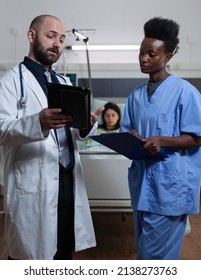 Doctor Reading Lab Results From Digital Tablet To Nurse Holding Clipboard With Medical History At Patient Bedside In Hospital Ward. Health Care Specialists Using Modern Technology For Clinical