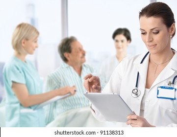 Doctor recording information to electronic tablet at patients bed, team working in background.? - Powered by Shutterstock