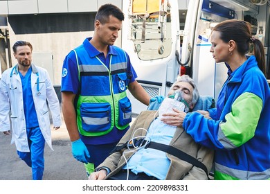 Doctor Rushing To Senior Patient With Breathing Mask On Ambulance Stretcher Who Was Brought By Paramedics In Ambulance. EMS