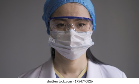 Doctor In Safety Glasses And Mask. Portrait Of Asian Woman In Medical Uniform And Goggles Looking At Camera On Gray Background.