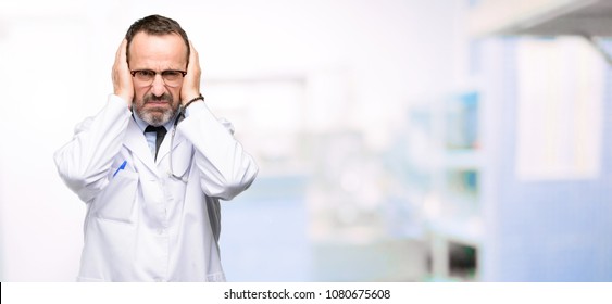 Doctor Senior Man, Medical Professional Covering Ears Ignoring Annoying Loud Noise, Plugs Ears To Avoid Hearing Sound. Noisy Music Is A Problem. At Hospital