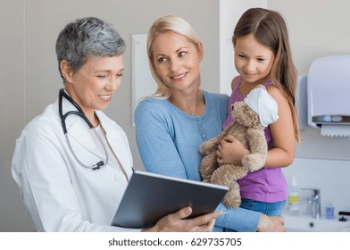 Doctor Showing Medical Report To Mother On A Digital Tablet In Hospital. Mother Carrying Daughter Looking At Pediatrician Holding Tablet. Little Girl With Teddy Bear Looking At Digital Tablet Screen.