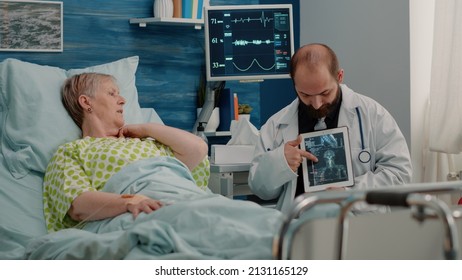 Doctor Showing Osteopathy X Ray On Tablet To Elder Woman In Bed At Nursing Home. Sick Patient Looking At Radiography Scan For Diagnosis And Healthcare Treatment With Help From Medic