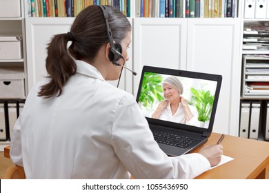 Doctor sitting at the desk of her office with headset and laptop, taking notes during a video call with a patient suffering from shoulder pain, telehealth or telemedicine concept