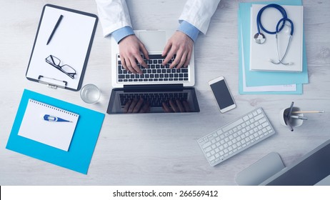 Doctor sitting at office desk   working his laptop and medical equipment all around  top view