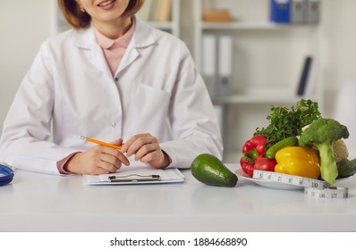 Doctor sitting at table with fresh raw fruit and vegetables and individual weight loss and slimming plan. Cropped female dietitian or nutritionist recommends sticking to diet and eating healthy food - Shutterstock ID 1884668890