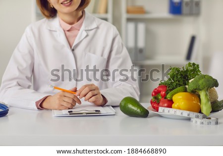 Doctor sitting at table with fresh raw fruit and vegetables and individual weight loss and slimming plan. Cropped female dietitian or nutritionist recommends sticking to diet and eating healthy food