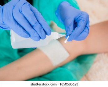 A doctor in sterile blue gloves processes a cotton swab with hydrogen peroxide to disinfect a wound on the patient's knee in the background.