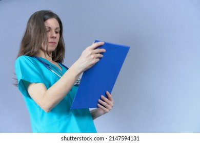 Doctor With Stethoscope Looking At Medical Report In Aquamarine Uniform Sideways From Below On Blue Background