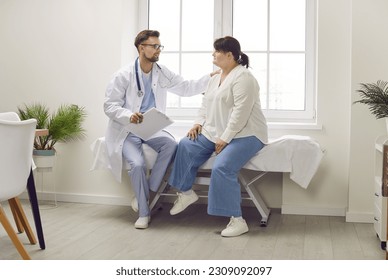 Doctor supporting patient. Man physician or cardiologist sitting on medical examination bed together with overweight woman, talking about treatment, holding hand on her shoulder and calming her down - Powered by Shutterstock