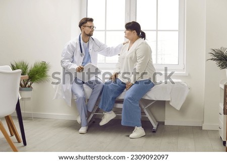 Doctor supporting patient. Man physician or cardiologist sitting on medical examination bed together with overweight woman, talking about treatment, holding hand on her shoulder and calming her down