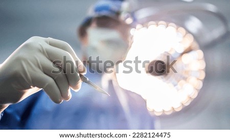 Doctor or surgeon in blue uniform holding surgical knife or scalpel to do surgery inside operating room in hospital under surgical lamp.People pick up surgical blade with white clean space.