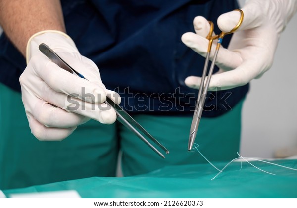 Doctor\
surgeon with disposable glove on hand holding forceps and scissor,\
stitch up wound or incision with suture thread over green fabric.\
Medical equipment of stainless tool for\
surgery.