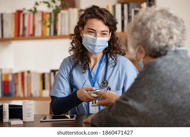 Doctor with surgical mask while visiting patient at home. Senior woman with doctor while doing coronavirus test and using oximeter. Rear view of old woman giving finger to doctor for oximeter analysis