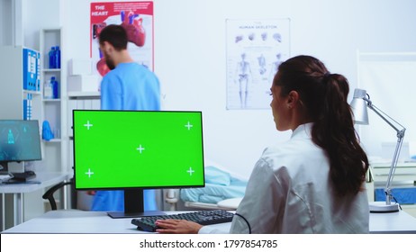 Doctor Taking Notes On Computer With Chroma Key Green Screen And Nurse In Blue Uniform In Hospital Cabinet. Medic In White Coat Working On Monitor With Blank Space In Clinic Cabinet To Check Patient