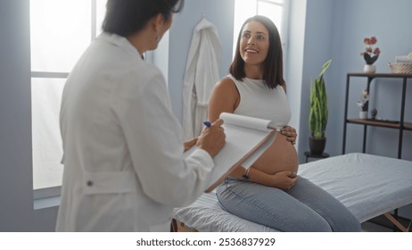 Doctor taking notes while pregnant woman sits on examination table in clinic room during medical appointment - Powered by Shutterstock