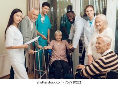 The Doctor Is Talking To An Elderly Woman In A Nursing Home. He Smiles. Nearby Are Medical Staff And Other Senior Men. They Are Looking At The Camera. She Is Dripping A Medical Dropper.