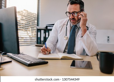 Doctor talking over his mobile phone and making notes in a book. Medicine professional talking on phone and writing in his diary. - Powered by Shutterstock