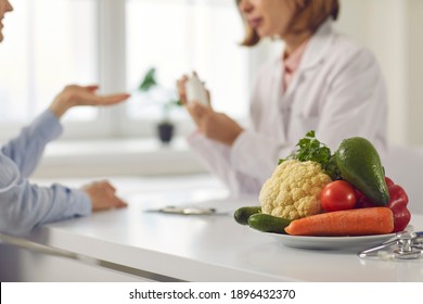 Doctor Talking To Patient And Teaching Her To Take Medicine, Eat Healthy Food And Stick To Diet. Nutrition Education And Disease Prevention Concept. Blurred Background, Bowl Of Vegetables In Close-up