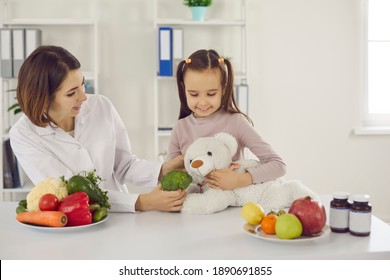 Doctor Teaching Child To Eat Fresh Healthy Food. Dietitian Or Nutritionist Talking To Little Girl, Playing Fun Game Of Feeding Teddy Bear And Telling About Benefits Of Eating Raw Fruit And Vegetables