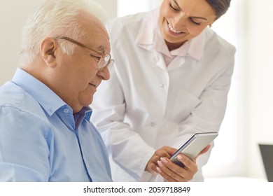 Doctor Teaching Senior Patient To Use Healthcare App For Setting Medication Reminder Or Tracking Health Indicators. Smiling Nurse In Assisted Living Facility Showing Photo Of Grandchildren To Old Man