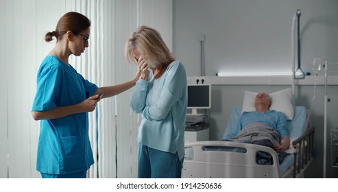 Doctor Telling Bad News To Aged Male Patient Wife In Hospital. Senior Lady Talking To Nurse And Crying Discussing Diagnosis And Treatment Of Sick Man Lying In Bed